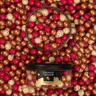 A clear plastic jar with a black lid filled with assorted gold, red and white speckled 'LAKRIDS BY BÚLOW' in cherry, caramel and raspberry flavours against a background filled with more of the gold, red and white speckled Lakrids confectionary.