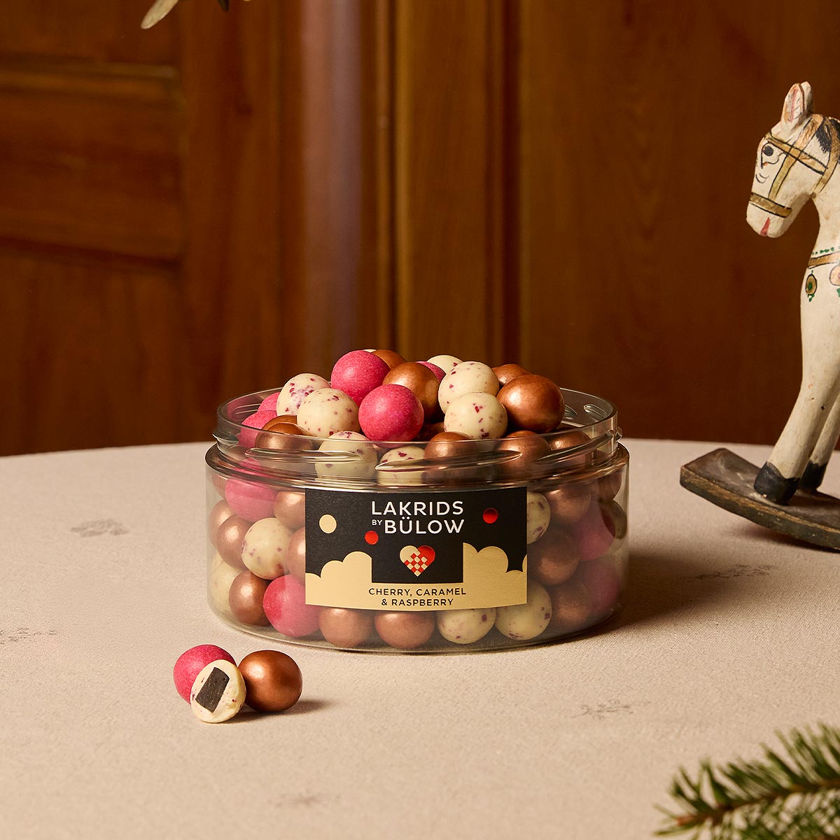An open clear plastic jar filled with assorted gold, red and white speckled 'LAKRIDS BY BÚLOW' in cherry, caramel and raspberry flavours against on a table with a cream tablecloth next to a sprig of fir and a rocking horse ornaments