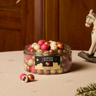 An open clear plastic jar filled with assorted gold, red and white speckled 'LAKRIDS BY BÚLOW' in cherry, caramel and raspberry flavours against on a table with a cream tablecloth next to a sprig of fir and a rocking horse ornaments