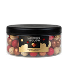 A clear plastic jar with a black lid filled with assorted gold, red and white speckled 'LAKRIDS BY BÚLOW' in cherry, caramel and raspberry flavours against a white background.