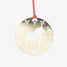A round, metallic gold Christmas ornament featuring a white silhouette of a Dala horse in the center, surrounded by decorative cutout patterns, hanging from a red string against a light gray background.