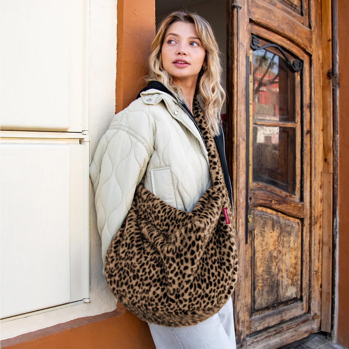 A woman wearing a cream quilted puffer jacket and white pants carries a large fuzzy leopard-print shoulder bag while leaning against a rustic wooden door.