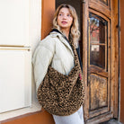 A woman wearing a cream quilted puffer jacket and white pants carries a large fuzzy leopard-print shoulder bag while leaning against a rustic wooden door.