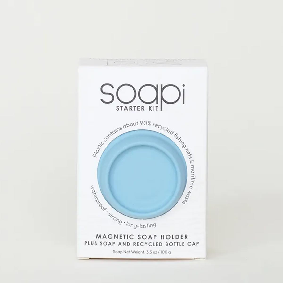A white rectangular box for the Soapi Starter Kit, featuring a circular cutout that reveals a light blue magnetic soap holder inside. The packaging includes text about recycled fishing nets and mentions the magnetic soap holder plus soap and recycled bottle cap.